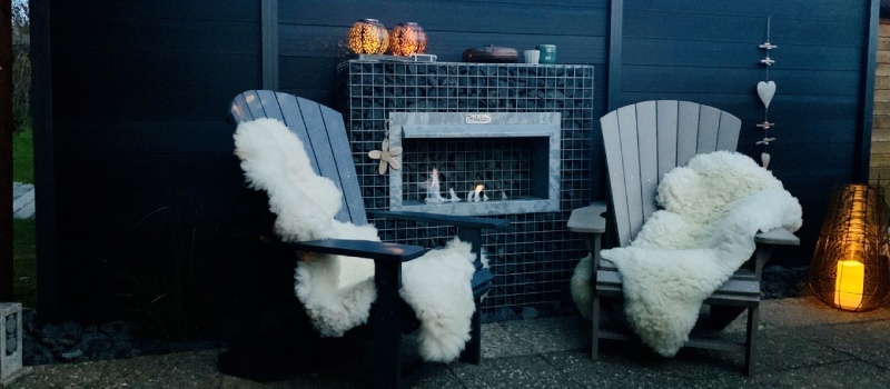 Geniet van warmte en sfeer in uw tuin met een bio ethanol tuinhaard Een sfeervolle bio ethanol haard verwerkt in een schanskorf, geplaatst tussen twee comfortabele stoelen met schapenvacht, in een moderne tuinsetting.